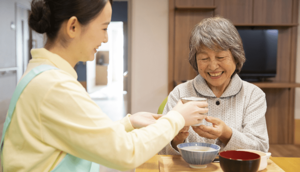 介護職員が入居者に食事を提供しており、コミュニケーションの様子が見られます。